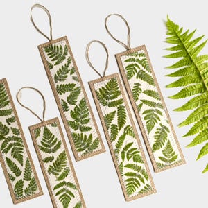 May include: Four rectangular bookmarks featuring a white background with a green fern leaf pattern. Each bookmark has a tan stitched border and a thin brown twine loop attached to the top. A single green fern frond is positioned to the right.