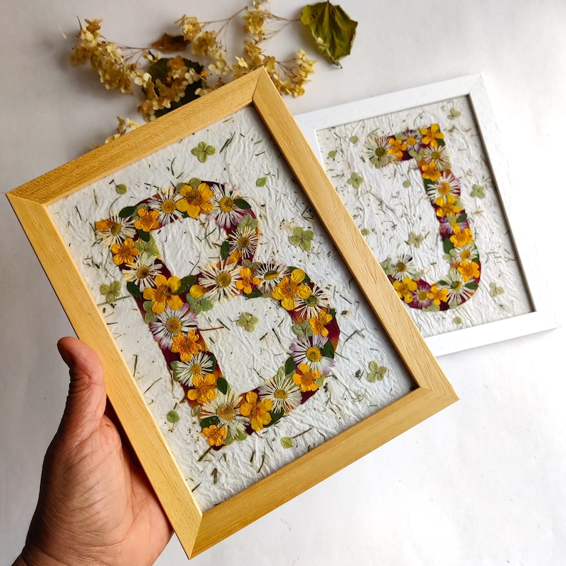 May include: Two rectangular frames containing pressed flower art on textured white paper. One frame is light wood and displays the letter B, while the other is white and displays the letter J. Both letters are formed by yellow and white flowers.