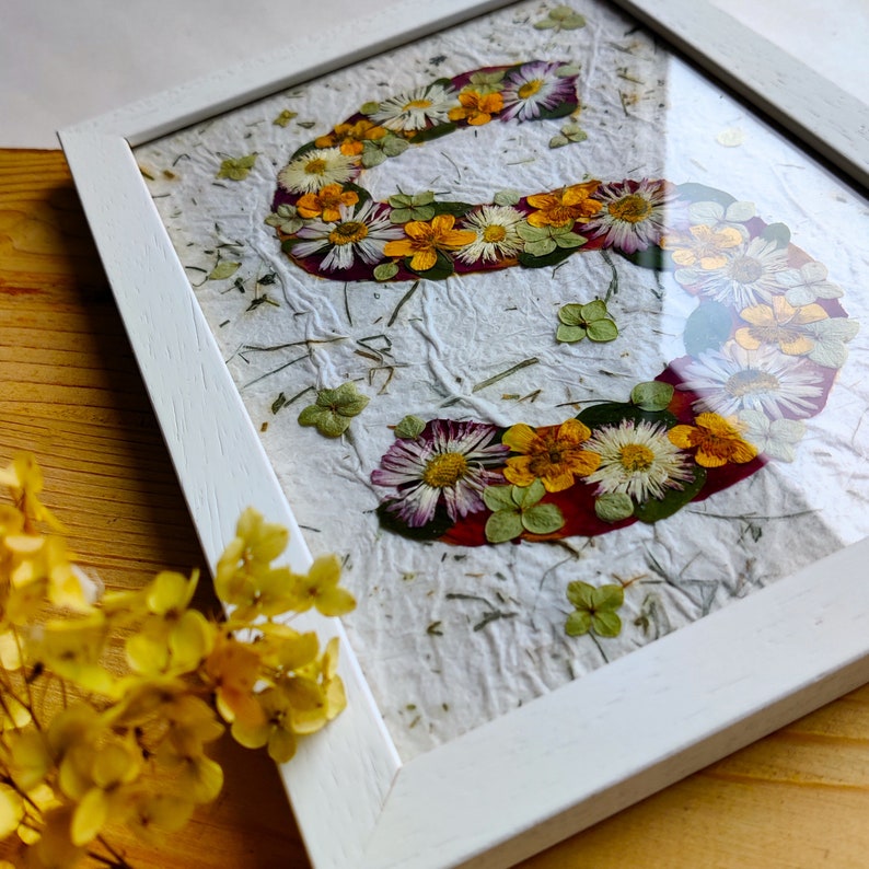 May include: A white wooden frame containing a piece of textured white paper with the letter S formed by pressed flowers. The floral arrangement includes white daisies, yellow buttercups, and small green leaves, all preserved under glass.