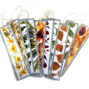 May include: A collection of eight rectangular bookmarks featuring pressed flowers and leaves encased in clear material with stitched grey edges and attached twine loops. Each bookmark displays unique botanical patterns including yellow flowers, green ferns, and autumn leaves.