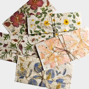 Könnte beinhalten: Eine Sammlung rechteckiger Papierumschläge mit eingearbeiteten gepressten Blumen und Blättern in den Farben Rot, Gelb, Grün, Rosa und Blau. Jeder Umschlag ist mit einer dünnen, naturfarbenen Schnur verschlossen, die zu einer Schleife gebunden ist.