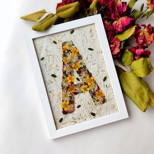 Könnte beinhalten: Ein weißer rechteckiger Rahmen mit einem strukturierten cremefarbenen Papier, auf dem der Buchstabe A aus gepressten gelben, weißen und violetten Blumen geformt ist. Kleine grüne Blätter sind auf der Papieroberfläche im Rahmen verteilt.