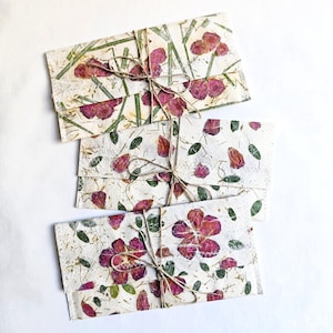 Könnte beinhalten: Drei handgefertigte Papierumschläge mit Blumen- und Blattmotiven. Die Umschläge sind mit Bindfaden gebunden. Das Papier hat einen strukturierten, cremefarbenen Hintergrund mit gepressten rosa Blüten und grünen Blättern.