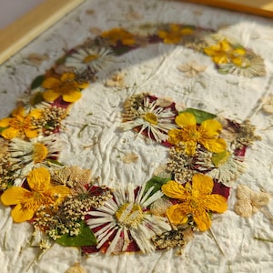 May include: A framed artwork featuring a letter G formed by pressed dried flowers. The composition includes bright yellow buttercups, white daisies, and small green leaves arranged on a textured, off-white handmade paper background.