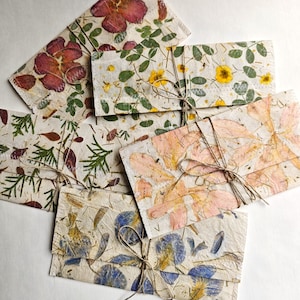 Op de afbeelding: Een verzameling rechthoekige handgemaakte papieren enveloppen met ingebedde gedroogde bloemen en bladeren in tinten rood, geel, roze en blauw. Elke envelop is vastgemaakt met een dun, natuurlijk bruin touwtje dat in een strik is geknoopt.