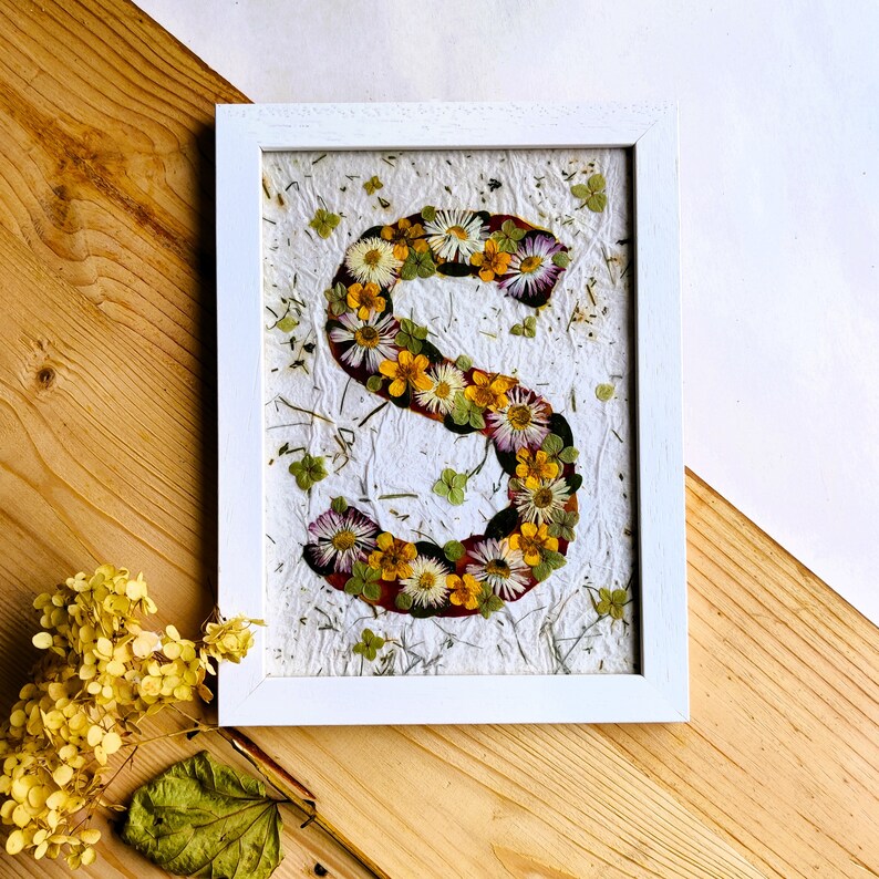 May include: A white rectangular frame containing a piece of textured white paper with the letter S formed by an arrangement of pressed dried flowers in shades of yellow, white, and purple, accented with small green leaves and scattered botanical bits.