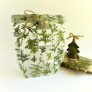 Può includere: Un sacchetto di carta bianca traslucida con un motivo di rami di conifere verdi, legato con un fiocco di spago marrone e un'etichetta regalo rettangolare abbinata. Accanto si trova un piccolo ornamento per albero in tessuto verde.