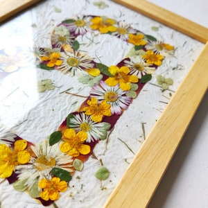 May include: A framed artwork featuring the letter P formed by pressed yellow buttercups, white daisies, and green leaves on a textured white paper background. The piece is enclosed in a light-colored wooden frame with a clear glass front cover.