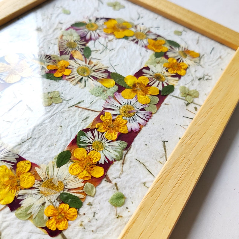 May include: A framed artwork featuring the letter P formed by pressed yellow buttercups, white daisies, and green leaves on a textured white paper background. The piece is enclosed in a light-colored wooden frame with a clear glass front cover.