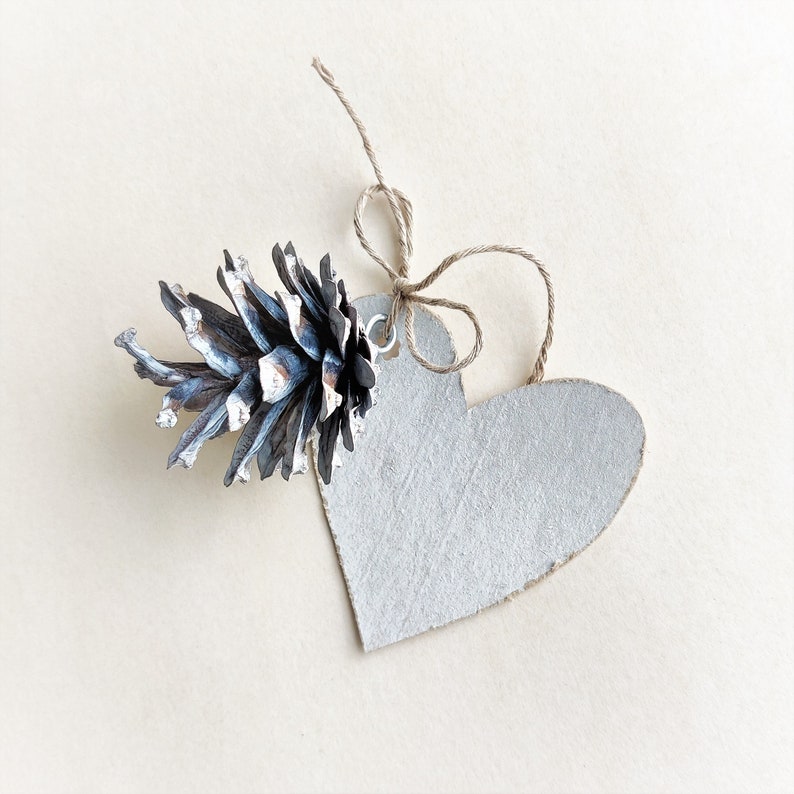 May include: A white heart-shaped tag with a brown twine bow and a small, dried pine cone attached.