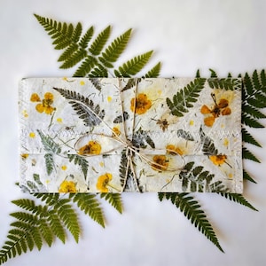 以下が含まれることがあります： 黄色い花と緑のシダの葉をあしらった、手作りの紙の封筒。封筒は紐で結ばれ、縁には白いステッチが施されています。緑のシダの葉が白い背景の封筒の周りに配置されています。