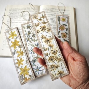May include: Four handmade bookmarks featuring pressed flowers. Each bookmark showcases a different floral design, including yellow flowers, blue flowers, and brown flowers. The bookmarks are crafted from a natural, beige material and have a decorative border.