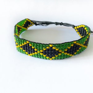 May include: A beaded bracelet with a pattern of green, yellow, and black beads. The bracelet has a woven design with a repeating geometric pattern. The bracelet is adjustable with black cords and is set against a white background.