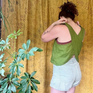 May include: A green linen tank top with a criss-cross back design. The tank top is worn over a brown tank top and paired with light gray corduroy shorts.