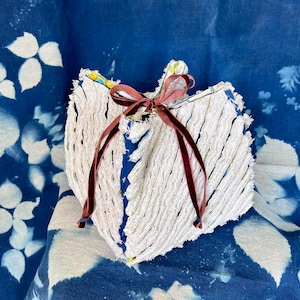 May include: A white, textured, heart-shaped pouch with a dark red ribbon closure. The pouch has a blue interior and is set against a blue background with white floral patterns. The pouch is approximately 6 inches wide.