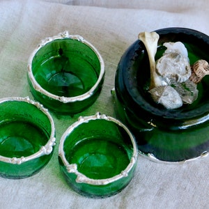 May include: A set of four green glass votive candle holders with silver-colored rims. One larger holder contains a bone and small stones. The candle holders are made from recycled glass bottles.