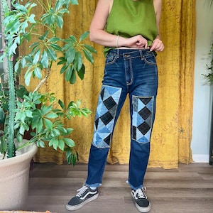May include: Dark wash denim jeans with patchwork knee details. The patches feature a geometric design in shades of blue and black. The jeans have a straight leg fit and a frayed hem.
