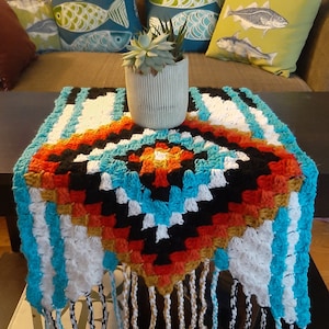 Puede incluir: Un camino de mesa de ganchillo con un patrón geométrico en tonos turquesa, blanco, negro, naranja y marrón. El camino de mesa tiene flecos en todos los lados y está sentado en una mesa de café con un libro y una suculenta en maceta.