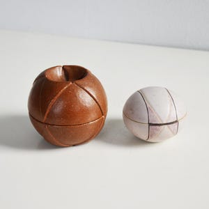 Puede incluir: Dos esferas decorativas de cerámica. Una es un recipiente esférico marrón más grande con un diseño tallado. La otra es una esfera blanca más pequeña con un patrón geométrico. Ambas están sobre una superficie blanca.