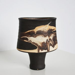 Puede incluir: Un jarrón de cerámica negro con la parte superior acampanada y una base de pedestal. El jarrón presenta un diseño único con patrones abstractos crema y marrón. El borde interior es de color marrón claro.