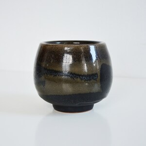May include: A small, dark brown ceramic cup with a glossy finish. The cup has a rounded shape and a slightly flared rim. The glaze has a mottled appearance, with shades of black and brown.
