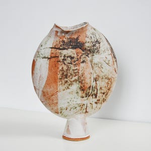 May include: A handcrafted ceramic vase with an unusual, organic form. The vase has a mix of colours, including white, orange, and brown, with a textured surface. The vase stands on a small, cylindrical base, enhancing its artistic appeal.