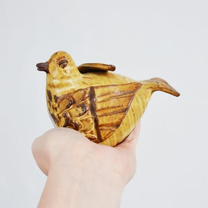 Könnte beinhalten: Keramik-Vogelfigur in Gelb- und Brauntönen. Der Vogel hat detaillierte Flügel- und Körpermuster, einen dunkelbraunen Schnabel und ein kleines Flügeldetaill auf dem Rücken. Die Figur wird in einer Hand vor weißem Hintergrund gehalten.