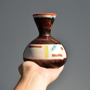 May include: A small, handcrafted ceramic vase with a dark brown speckled body and a white band featuring colorful geometric shapes. The vase has a narrow neck and a flared rim with orange and blue accents.