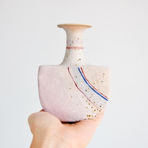 May include: A small, white ceramic vase with a pink glaze and a unique shape. It has a narrow neck and a wide base, and is decorated with thin red, blue, and white lines.