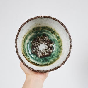 Puede incluir: Un cuenco de cerámica blanca con un esmalte verde y marrón. El cuenco tiene un diseño decorativo de una flor en el centro.