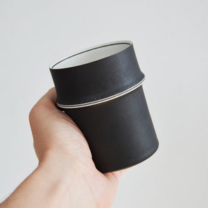 Puede incluir: Una taza de cerámica negra con un borde blanco y dos finas líneas negras alrededor de la parte superior.