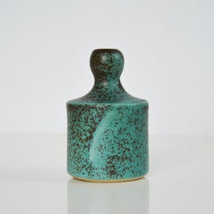 May include: A small, teal-colored ceramic vase with brown speckles. The vase has a rounded, bulbous top and a cylindrical base.