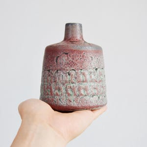 Puede incluir: Un jarrón de cerámica texturizada en tonos burdeos, grises y verdes. El jarrón tiene un cuerpo cilíndrico con un patrón texturizado y un cuello estrecho. El jarrón se sostiene en una mano sobre un fondo blanco. Mide unos 15 cm de alto.