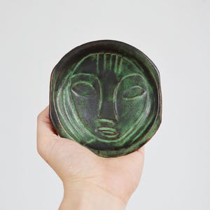 May include: A round ceramic dish featuring a dark green and black glaze with an embossed, stylised human face design. The surface has a smooth, glossy finish with deep green highlights accentuating the facial features and the circular rim.