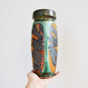 May include: A cylindrical ceramic vase featuring a dark green rim and base with a vertical green stripe separating two panels of textured orange clay. The surface is decorated with bold, abstract black brushstroke patterns across the orange sections.