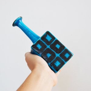 May include: A blue ceramic vase with a geometric pattern of black squares. The vase has a square base and a tall, thin neck.