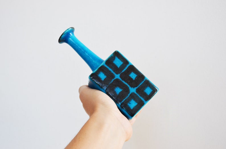 K&ouml;nnte beinhalten: Eine blaue Keramikvase mit einem geometrischen Muster aus schwarzen Quadraten. Die Vase hat einen quadratischen Boden und einen hohen, d&uuml;nnen Hals.