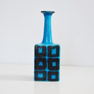 K&ouml;nnte beinhalten: Eine kleine, blaue Keramikvase mit einem geometrischen Muster aus schwarzen Quadraten. Die Vase hat einen schmalen Hals und einen quadratischen Boden.