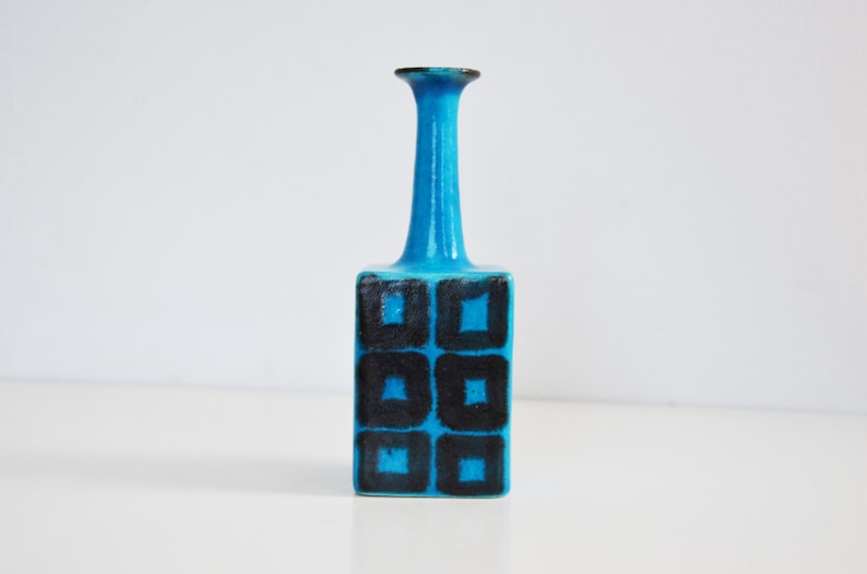 K&ouml;nnte beinhalten: Eine kleine, blaue Keramikvase mit einem geometrischen Muster aus schwarzen Quadraten. Die Vase hat einen schmalen Hals und einen quadratischen Boden.