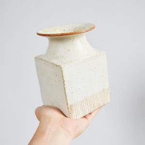 Puede incluir: Un jarrón de cerámica de color crema con base cuadrada y parte superior redondeada. El jarrón tiene una superficie texturizada con una banda de líneas verticales cerca de la base y un borde marrón. El jarrón está en la mano de una persona.