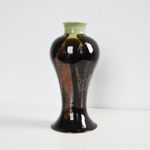 May include: A ceramic vase with a flared base and a narrow neck. The body features a glossy dark brown glaze with a textured, mottled copper-coloured section. The rim and neck are finished in a smooth, pale light green glaze with a reflective surface.
