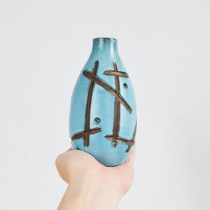 Puede incluir: Un jarrón de cerámica azul claro con cuello estrecho y cuerpo redondeado, que presenta líneas oscuras entrecruzadas pintadas a mano y pequeños acentos circulares en la superficie, presentado con un acabado esmaltado suave y liso.