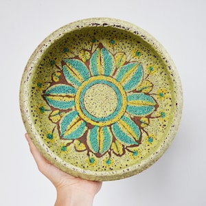 Peut inclure: Un grand bol rond en céramique avec un motif floral vert, jaune et bleu. Le bol a un glaçage moucheté et un bord légèrement surélevé.