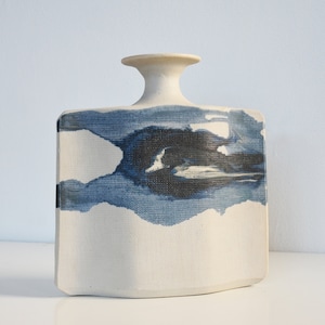 Op de afbeelding: Een witte keramische vaas met een vierkant lichaam en een smalle hals. De vaas heeft een blauw en zwart abstract ontwerp dat op de voorkant is geschilderd.