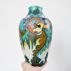 May include: A decorative vase with a wide body and a narrow neck. The vase features a colourful floral design in shades of teal, orange, yellow, and blue. The hand-painted details include stylised leaves and flowers, creating an artistic and elegant look.