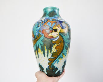 Puede incluir: Un jarrón decorativo con un cuerpo ancho y un cuello estrecho. El jarrón presenta un diseño floral colorido en tonos de verde azulado, naranja, amarillo y azul. Los detalles pintados a mano incluyen hojas y flores estilizadas, creando un aspecto artístico y elegante.