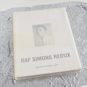 May include: A book titled "RAF SIMONS REDUX" with a portrait on the cover. The book is on a silver, mesh-like surface. The book's cover is off-white with gray text and a black and white portrait.