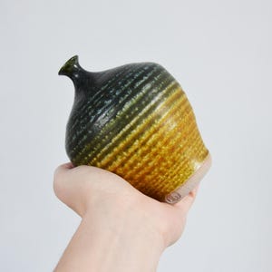 Könnte beinhalten: Eine handgefertigte Keramikvase mit einer einzigartigen Form und strukturierter Oberfläche. Die Vase hat einen Farbverlauf, der von Dunkelgrün zu Goldgelb übergeht. Die Vase hat einen kleinen Ausguss.