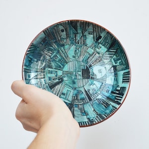 May include: A teal ceramic bowl with a geometric design of black and white lines and shapes. The bowl has a brown rim.