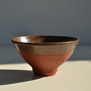 May include: A small, brown ceramic bowl with a dark brown glaze on the top half and a lighter brown glaze on the bottom half. The bowl has a slightly flared rim and a rounded base.